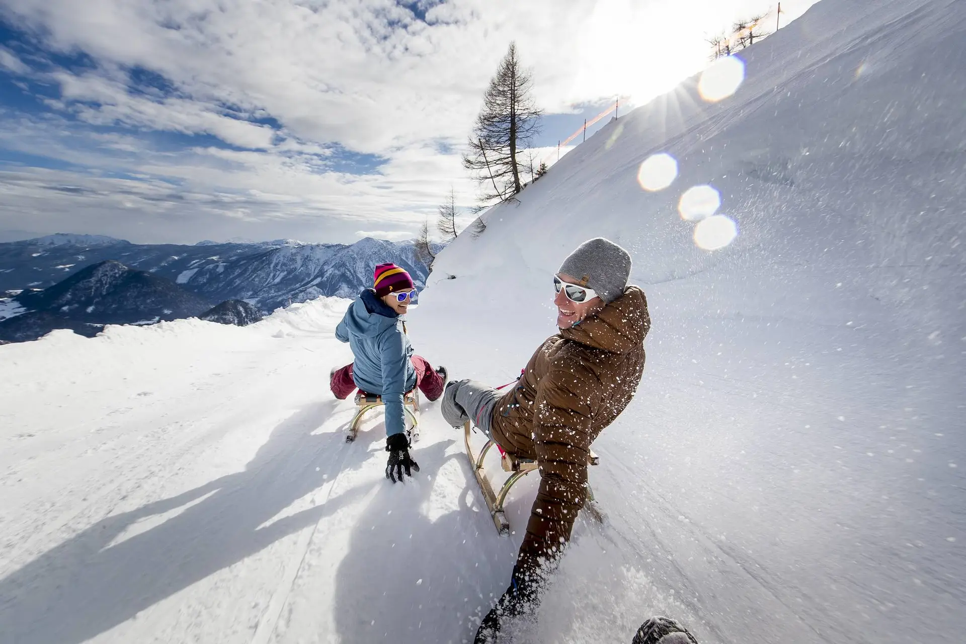 Zwei Personen rodeln an einem sonnigen Tag einen schneebedeckten Hang hinunter. Die Gesichter sind fröhlich, im Hintergrund sind eine Bergkette und ein teilweise bewölkter Himmel zu sehen.